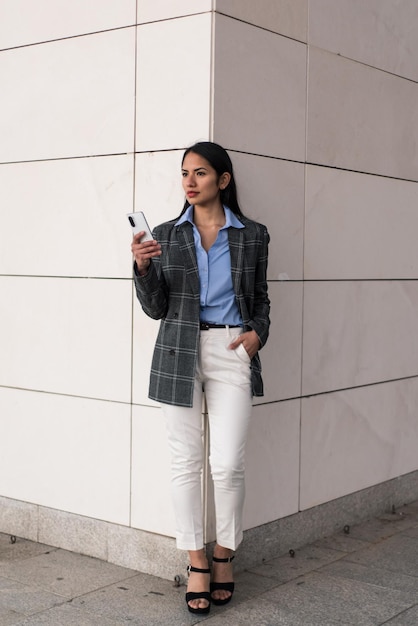 Фото Женщина в клетчатом блейзере и белых брюках стоит у стены, прислонившись к стене, держа телефон.
