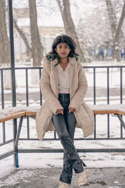 Фото Женщина в коричневом пальто и черных брюках сидит на скамейке в снегу