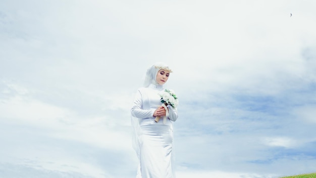 사진 하얀 드레스를 입은 한 여성이 푸른 하늘 앞에서 꽃을 들고 있습니다.