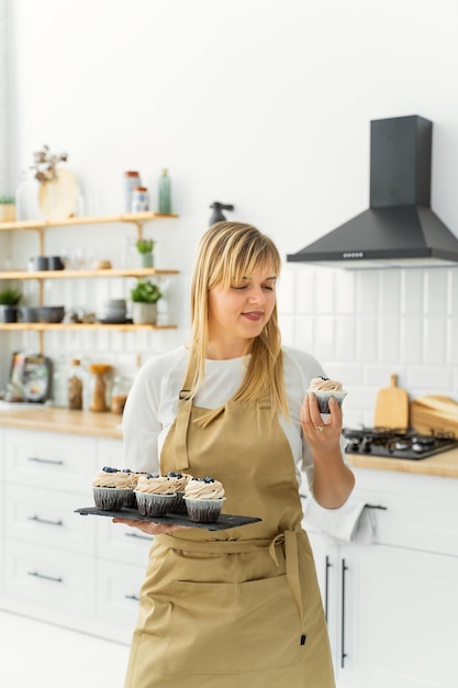 Фото Женщина в фартуке держит на кухне поднос с кексами.