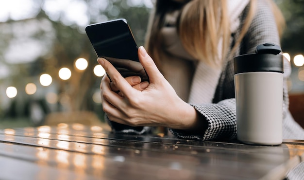 写真 秋のコートとスカーフを着た女性が、通りのカフェで携帯電話を手に持っています。閉じる