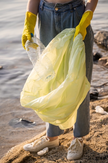 노란색 고무장갑을 끼고 플라스틱 쓰레기 봉투를 든 여성이 호숫가에 서 있습니다. 자연과 저수지를 청소합니다.