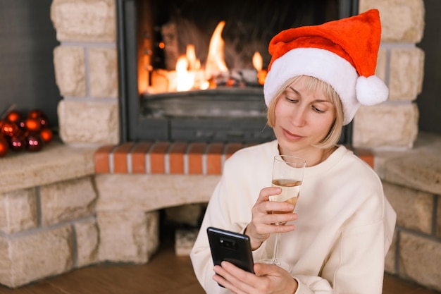 Женщина в шляпе деда мороза общается по телефону у горящего камина в уютном доме