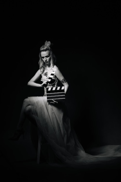 사진 한 여성이 흑백 클래퍼보드와 함께 사진을 찍기 위해 포즈를 취하고 있습니다. 이 이미지는 빈티지하고 고전적인 느낌을 가지고 있습니다.
