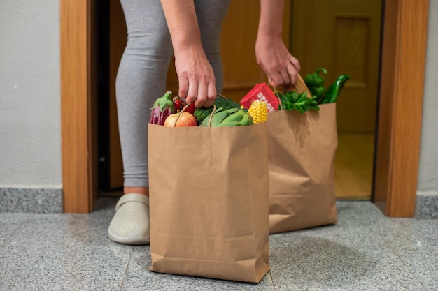 写真 女性が家のドアで食べ物の袋を手に取るコロナウイルス検疫家にいるオンラインショッピング検疫と自己隔離の間に野菜と果物の配達