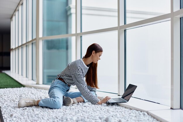 写真 女性はオフィスのロビーの大きな窓のそばに座り、インターネットを介してオンラインでノートパソコンを操作し、世界中のどこでもフリーランスの仕事をしています。