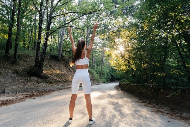 夕焼けの鬱蒼とした森の道でトレーニングを実行する前に、若い美しい少女が背中を向けてポーズをとる健康的なライフスタイルと新鮮な空気の中で走る