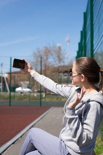 写真 スポーツウェアを着た若いブロンドの女性が、スタジアムのベンチに座ってビデオ通話をしているスポーツをした後、休んでいる