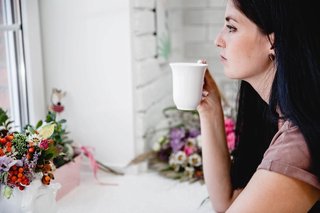 写真 長い髪の若い黒髪のヨーロッパの女性は、花のあるテーブルに座って、wからコーヒーを飲みます...