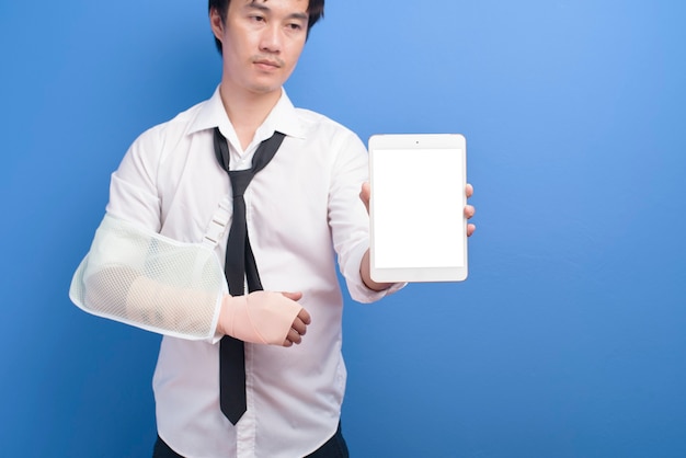 Молодой бизнесмен с травмированной рукой в перевязке, используя планшет на синем фоне в студии, страховании и концепции здравоохранения