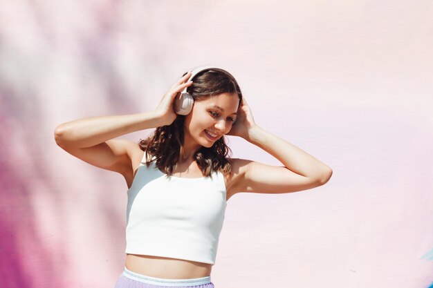 Молодая возбужденная женщина в топе и юбке в наушниках слушает музыку, ходит, танцует с поднятыми руками на открытом воздухе у розовой стены