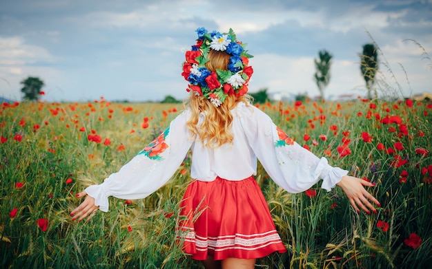 写真 明るい民族衣装を着て、頭に花輪を捧げ、腕を広げている少女が、赤いポピーの畑を歩いています。