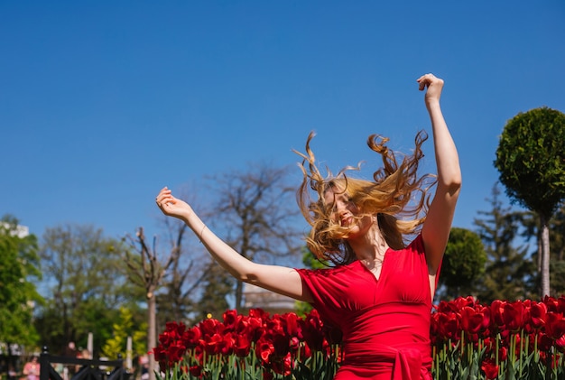 Фото Молодая девушка сидит на солнышке среди тюльпанов. в красном платье ее волосы развевались на ветру. атмосфера свободы и летнего настроения.