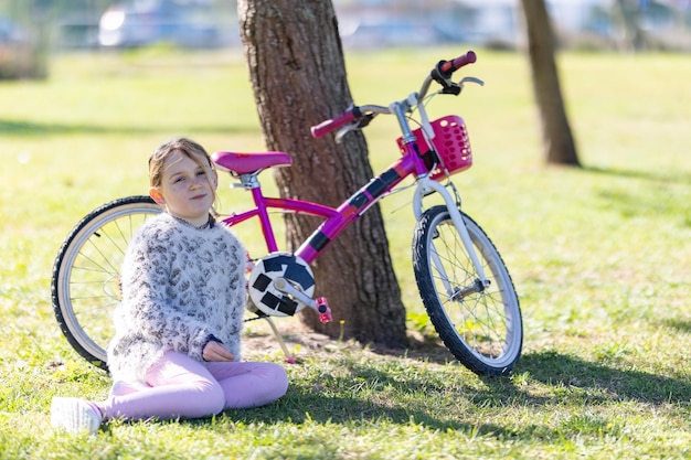 写真 若い女の子がピンクの自転車の隣の草の上に座っている