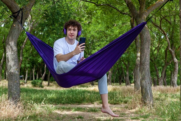 写真 若い男が紫色のハマックに座ってヘッドフォンで音楽を聴いて自然を楽しんでいます