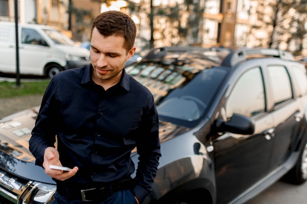 車の近くの通りに立っている若い男や暗いシャツを着たビジネスマンがスマートフォンを見ます。運転手は乗客またはクライアントを待っています。都市交通のコンセプト