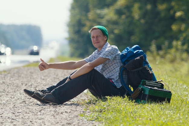 写真 道路のそばに座って、車のヒッチハイクをキャッチする若い男がクローズアップ