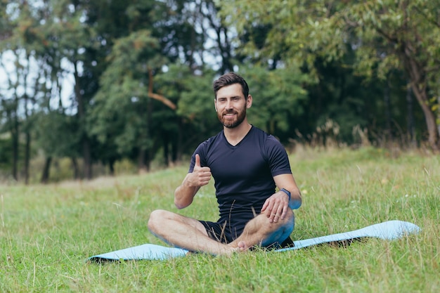 写真 マットの上に公園に座っているあごひげを生やした若い男性が瞑想し、呼吸、アクティブなライフスタイルを改善するための運動を行います