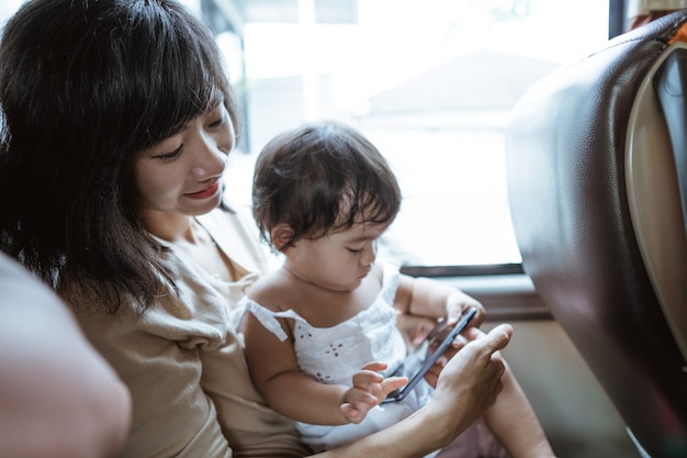 若い母親と少女が旅行中にバスに座っている彼らの携帯電話でビデオを見る