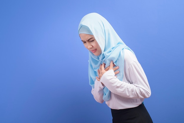 파란색 벽, 심장 마비 및 의료 개념을 통해 가슴 통증으로 고통받는 Hijab를 착용하는 젊은 무슬림 여성.