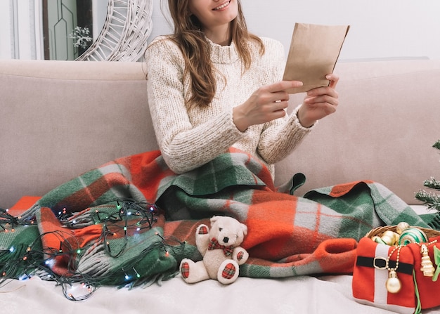 Фото Молодая симпатичная женщина в легком свитере сидит на кровати с елочными украшениями и читает письмо