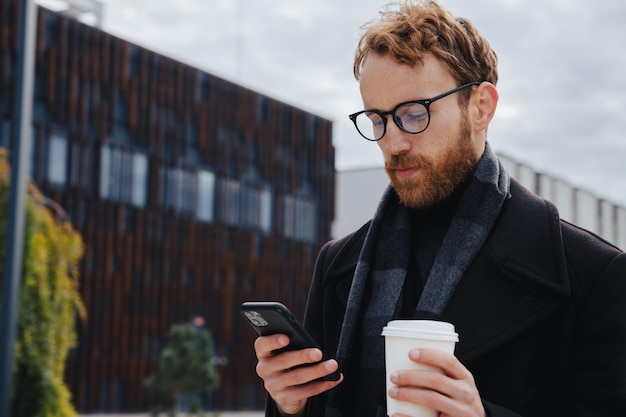 写真 都会的なスタイルを背景に眼鏡をかけた若い赤髪の男 スマートフォンでメールをチェックするビジネスマン メッセンジャーで通信し、コーヒーを飲む オンライン通信