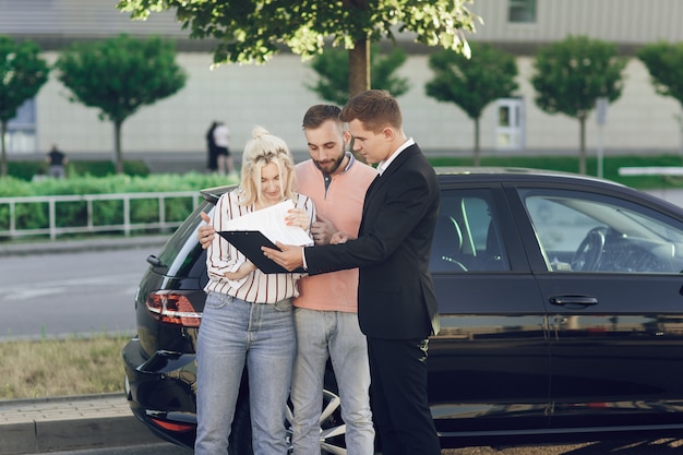 写真 若いセールスマンが新しい車を顧客に見せます。幸せなカップル、男性と女性が新しい車を購入します。若い人たちは車を買うために書類に署名します。