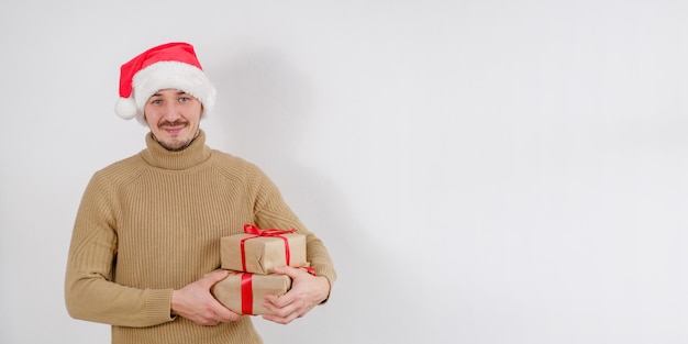 산타클로스 모자를 쓰고 선물 상자를 들고 웃고 있는 젊은 백인 남자가 격리된 흰색 배경에서 카메라를 쳐다본다
