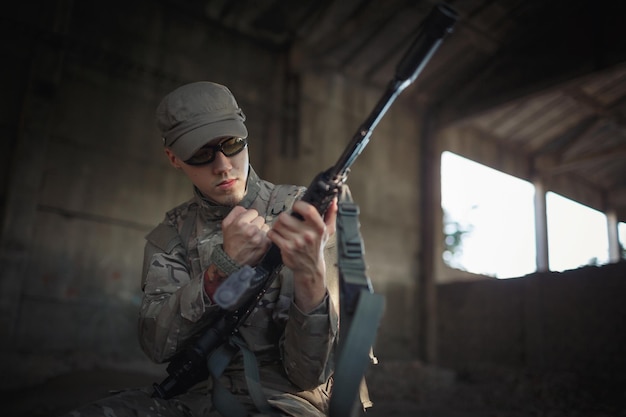 写真 キャップと戦術的な黒いゴーグルを身に着けている灰色のカモフラージュの制服を着た若いウクライナの兵士は、大きな破壊された建物内のコンクリートの床に座って、機関銃を掃除します