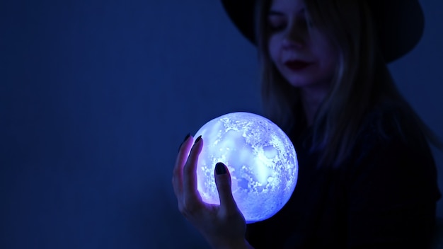 Фото Гадалка молодая женщина в шляпе держит волшебный шар. ярко-синий шар светится в темноте.
