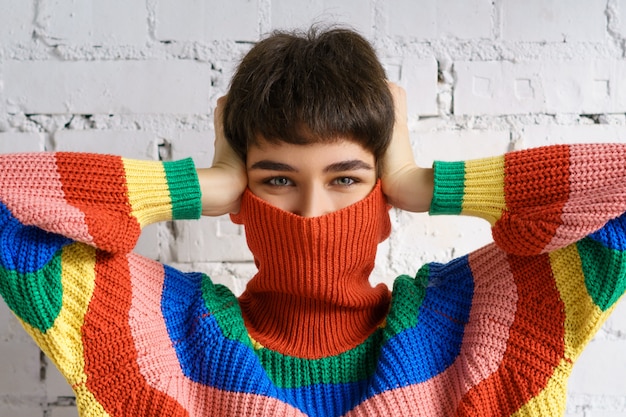 밝은 여러 가지 빛깔의 무지개 스웨터에 젊은 여자는 그녀의 얼굴을 숨기고 그녀의 손으로 그녀의 귀를 다루고 있습니다.