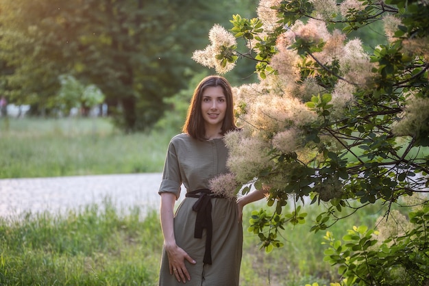 写真 緑のカーキ色のドレスを着た若い女性が公園で日没時にバラの茂みの煙のようなスカンピアのそばに立っています