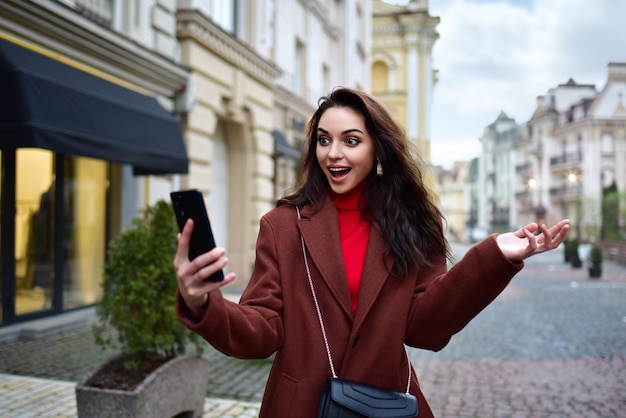 写真 赤いコートを着た若い女性が通りを歩いて 手に持ったスマートフォンを見て 喜びの感情を経験します