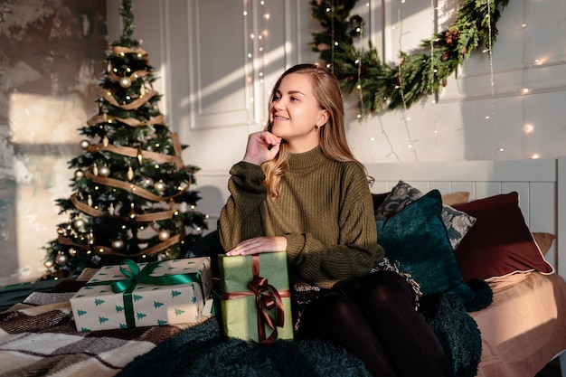 스웨터를 입은 젊은 여성이 크리스마스 트리를 배경으로 크리스마스로 장식된 침실에서 크리스마스 선물을 엽니다.