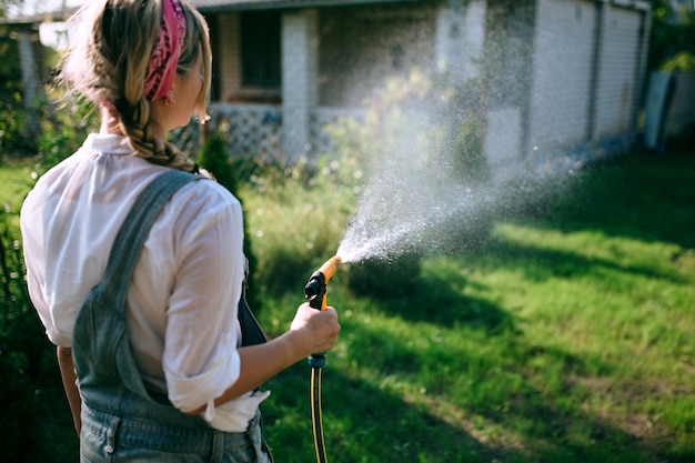 Фото Молодая женщина в белой рубашке и джинсовом комбинезоне поливает лужайку. концепция озеленения и ландшафтный дизайн