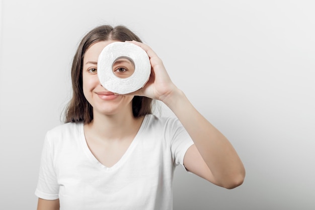 Фото Молодая женщина в белой футболке держит перед глазами рулон туалетной бумаги на сером фоне