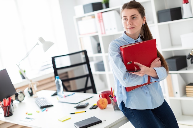 Молодая женщина в офисе стоит, опираясь на стол, держит телефон и папку с документами.