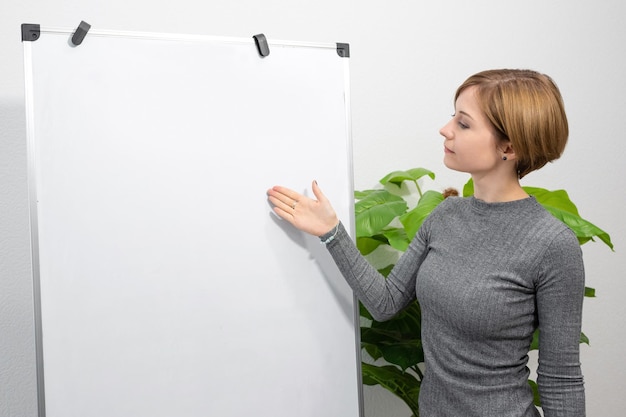사진 한 젊은 여성이 빈 플립 차트를 가리키는 밝은 방에 서 있습니다. 온라인 교사 교사