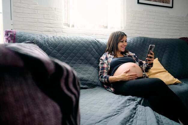 Фото Молодая женщина с очень поздней беременностью сидит на диване, смотрит и разговаривает по мобильному телефону.