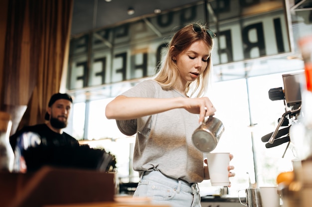 На фотографии изображена юная худощавая блондинка в повседневной одежде, добавляющая молоко в кофе в уютной кофейне. .