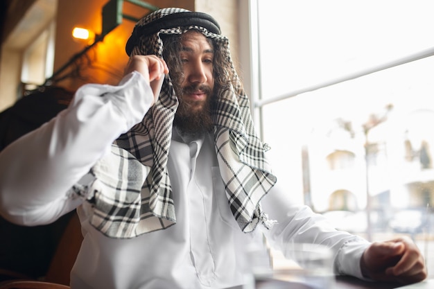 Aan de telefoon. Arabische zakenman die op kantoor werkt, zakencentrum met apparaat, gadget. Moderne Saoedische levensstijl. Man in traditionele kleding en sjaal ziet er zelfverzekerd, druk, knap uit. etniciteit, financiën