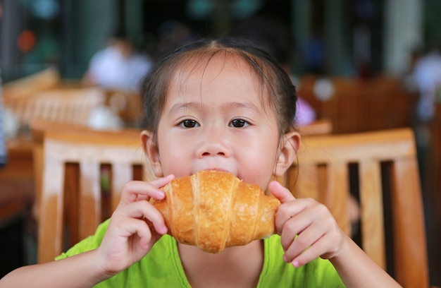 Aanbiddelijk Aziatisch kindmeisje die een beet van croissant in de ochtend nemen bij restaurant.