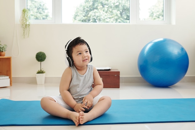 Aanbiddelijk glimlachend klein kind van gemengd ras dat op een yogamat zit en naar muziek luistert in een koptelefoon