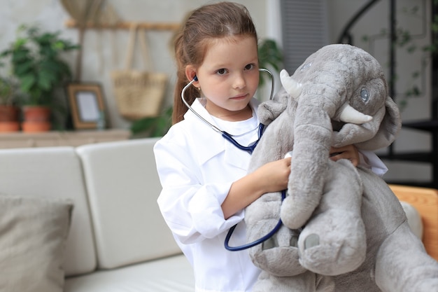 Aanbiddelijk kind verkleed als dokter die met speelgoedolifant speelt en zijn adem controleert met een stethoscoop.