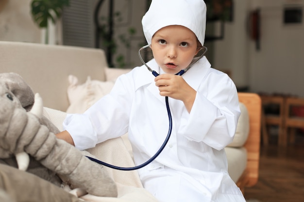 Aanbiddelijk kind verkleed als dokter die met speelgoedolifant speelt en zijn adem controleert met een stethoscoop.