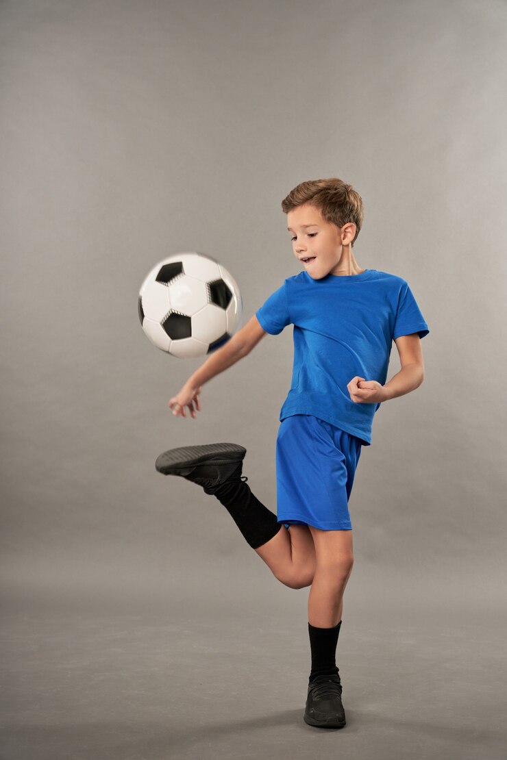 Aanbiddelijk mannelijk kind in korte broek die voetbaltrucs doet tegen een grijze achtergrond