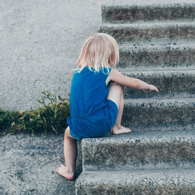 Aanbiddelijk meisje dat op aard lacht - gelukkig meisje in een blauwe jurk en door de knieën geslagen zit op de trap. Gelukkige jeugd.