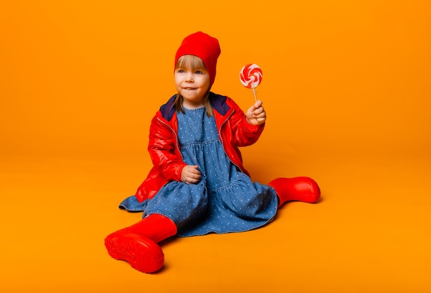 Aanbiddelijk meisje in een rood jasje en laarzen die een lolly op een gele achtergrond houden. herfst begrip.