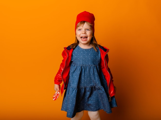 Aanbiddelijk meisje in een rood jasje en laarzen die een lolly op een gele achtergrond houden. herfst begrip.