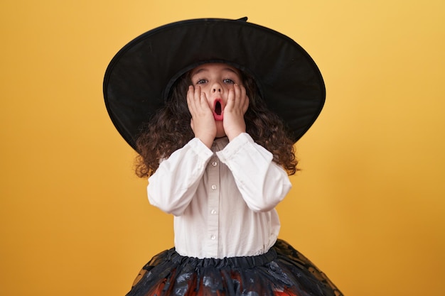 Aanbiddelijk Spaans meisje dat Halloween-kostuum draagt dat zich met verrassingsuitdrukking over geïsoleerde gele achtergrond bevindt
